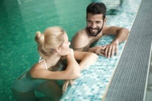 A man and woman in swimsuits are smiling and talking while resting at the edge of an indoor swimming pool.