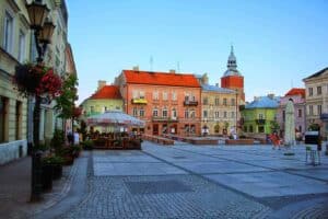 Town square with colorful historic buildings, outdoor caf&eacute; seating under umbrellas, people walking, and a tall church tower in the background&mdash;one of the best places near Ł&oacute;dź for a charming day trip.