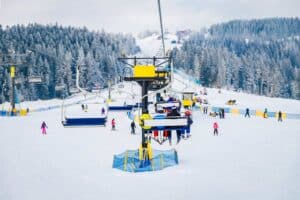 Skiers ride a chairlift at a snowy ski resort, with people skiing and snowboarding on the slopes surrounded by pine trees.