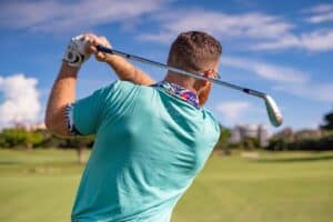 A man in a turquoise shirt swings a golf club on one of the best golf courses, bathed in sunlight and facing away from the camera.