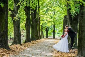 A bride and groom share a kiss on a bench along a tree-lined path covered with fallen leaves in a park, capturing the charm of romantic places in Poland.