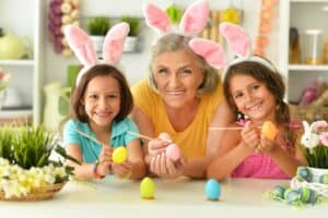 An older woman and two girls wearing bunny ears decorate Easter eggs at a table with plants and festive decorations, celebrating Polish Easter traditions.