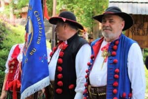Three people in traditional Polish folk costumes stand outdoors; one holds a blue flag with text, and greenery is visible in the background.