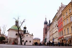 A city square with a small domed church, colorful historic buildings, and a tall Gothic tower in the background under a cloudy sky showcases some pros and cons of living in Poland as people stroll along the cobblestone street.