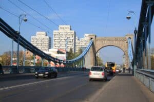 Suspension bridge with cars and pedestrians, blue cables, and stone arch; city buildings in the background under clear sky&mdash;a picturesque spot for those driving in Poland or visitors with a US license seeking scenic views.