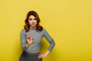 Woman with brown hair stands against a yellow background, holding one hand up in a stop gesture and her other hand on her hip, with a skeptical facial expression.