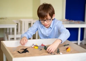 A young boy wearing glasses plays with toy dinosaurs in a sandbox, engaging in sand tray therapy.