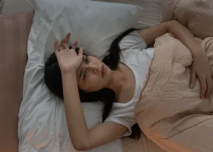 A woman in bed, hands on her head, contemplating her struggles with sleep and depression.