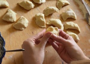 Hands shaping pierogi on a wooden surface, with several uncooked dumplings and a fork visible nearby&mdash;just like Grandma's recipe.