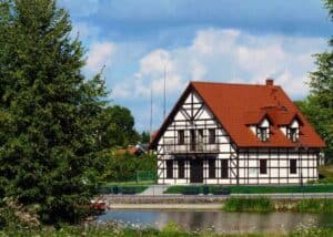 A white half-timbered house with a red roof stands by a pond, surrounded by trees and greenery under a partly cloudy sky&mdash;an idyllic scene that makes Mazury, Poland one of the best places to visit for travel enthusiasts.