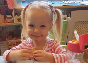 A young girl with pigtails and a crumb-covered face smiling at a table with cookies and a sippy cup, enjoying some of the best Polish food for kids.