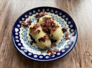 Three potato dumplings, made following a traditional Kartacze Recipe, are topped with diced, fried bacon and served on a patterned ceramic plate placed on a wooden surface.