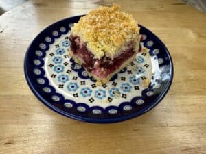A square piece of berry crumb cake, inspired by a classic Polish fruit cake, sits on a decorative plate with blue flower patterns, placed on a wooden table.