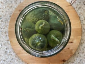 Overhead view of green figs submerged in liquid inside a clear glass jar, reminiscent of the traditional Ogorki Malosolne recipe, placed on a wooden surface.