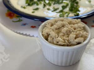 A small white ramekin filled with a beige spread sits on a tablecloth, embodying a classic Polish touch. In the background, a plate with a creamy dish garnished with chopped greens is partially visible, reminiscent of traditional Chrzan or horseradish recipe delicacies.