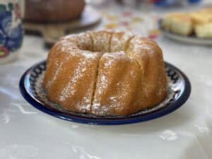 A round Polish Bundt Cake with a dusting of powdered sugar sits elegantly on a patterned plate, placed on a white tablecloth.