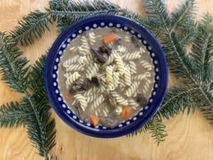 A bowl of pasta soup with fusilli, vegetables, and pieces of meat, placed on a wooden surface and surrounded by pine branches, reminiscent of a comforting Zupa Grzybowa recipe.