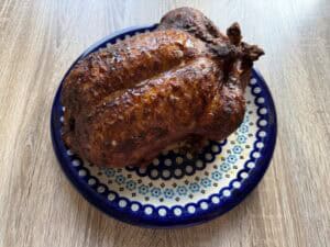 A whole roasted chicken with crispy, browned skin sits on a patterned blue and white plate on a wooden surface, reminiscent of a classic Polish duck recipe presentation.