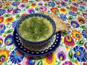 A blue bowl of Polish Dill Soup garnished with fresh dill sits elegantly on a matching saucer, accompanied by a slice of brown bread on a vibrant, floral-patterned tablecloth.
