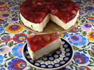 A slice of Polish cold cheesecake with strawberry jelly on a patterned plate sits in front of the remaining cake on a floral tablecloth.