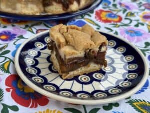 A square piece of layered pastry with a fruit filling, reminiscent of Polish Plum Cake, sits on a patterned plate atop a colorful floral tablecloth.