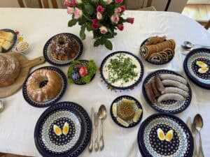 The table is set for a Traditional Polish Easter, featuring sausages, a bundt cake, sliced rolls, deviled eggs, and bread. A charming flower centerpiece sits on the white tablecloth with patterned plates.