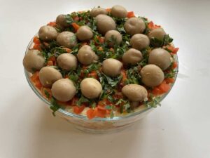 A glass bowl brimming with Polish forest glade salad reveals a layered marvel, topped with whole mushrooms, chopped red bell peppers, and parsley.
