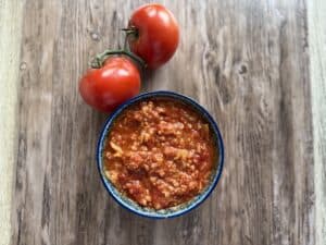 A bowl of tomato-based Polish stuffed cabbage soup sits on a wooden surface next to two fresh tomatoes still attached to the stem.