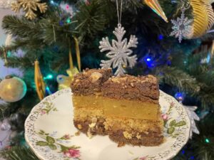 A floral plate holds two square pieces of Walnut Layered Cake in front of a decorated Christmas tree with gold and silver ornaments.