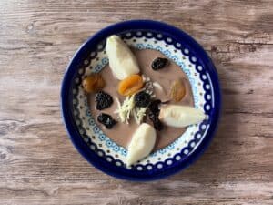 A patterned blue plate holds poached pears, dried apricots, prunes, shredded cheese, and chocolate sauce&mdash;an elegant spread reminiscent of desserts enjoyed after savoring traditional Polish duck blood soup.