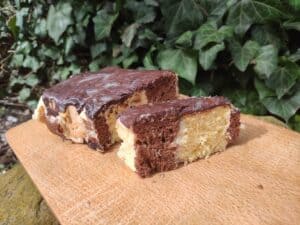 A sliced marble cake with chocolate and vanilla layers is placed on a wooden cutting board, set against a background of green ivy leaves. This delightful treat is reminiscent of the traditional Polish Meter-Long Cake, or Metrowiec, adding an elegant touch to any occasion.