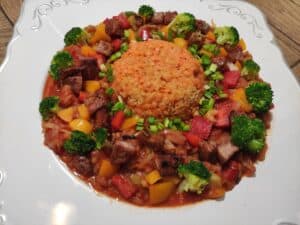A plated dish featuring a mound of rice surrounded by chunks of kielbasa, bell peppers, and broccoli, all served in a tomato-based sauce and garnished with chopped green onions.
