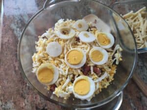 A glass bowl containing a mixture of shredded yellow and white cheeses, sliced hard-boiled Polish eggs, and small pieces of kielbasa.