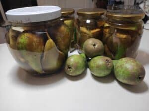 Jars filled with Grzuszki W Occie (Polish Vinegar Pickled Pears) and several fresh green pears placed in front of them on a white countertop.