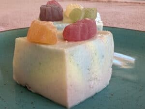 A square piece of pastel rainbow mousse, reminiscent of a No-Bake Rainbow Cake, is topped with colorful gummy bears on a green ceramic plate.