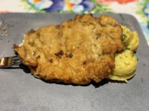 A breaded schnitzel, reminiscent of traditional Polish pork cutlets, sits on a plate alongside two scoops of herb-garnished mashed potatoes, all set against a floral tablecloth.