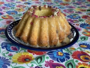 A White Chocolate Babka with glaze and pink sprinkles sits elegantly on a decorative plate, resting on a vibrant flower-patterned tablecloth.