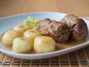A traditional Rolada Śląska sits on a bamboo mat, featuring two hearty meat rolls paired with boiled potatoes and cabbage, all drizzled with rich gravy&mdash;a classic recipe embodying comfort and flavor.