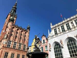 A bronze statue stands on a fountain in front of historic brick and white buildings under a clear blue sky.