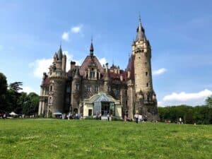 A large stone castle with multiple towers and pointed roofs stands on a grassy lawn under a blue sky, with people gathered around the entrance&mdash;one of the best day trips from Krakow for history and architecture enthusiasts.