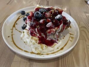 A plate with stacked pancakes topped with whipped cream, berry compote, blueberries, strawberries, powdered sugar, and a drizzle of caramel sauce.