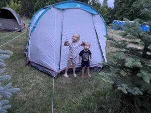 Two young children stand outside a gray tent on grass, surrounded by trees and other tents at one of the scenic Poland campsites, showcasing the joys of camping in Poland.