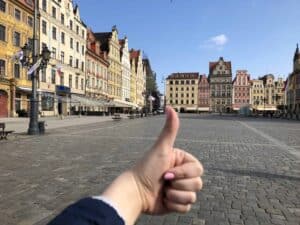 A hand giving a thumbs up in the foreground with a view of an empty European town square lined with colorful historic buildings, reflecting the impression that Wroclaw travel is enjoyable and highlights Wroclaw safety for visitors.