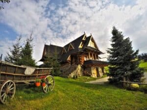 A rustic wooden house with a steep roof sits on a grassy lawn, next to an old wooden cart and pine trees, under a partly cloudy sky.