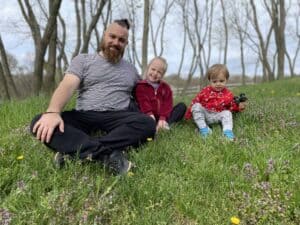 An adult and two young children sit on the grass in a park with trees in the background, all facing the camera and smiling.