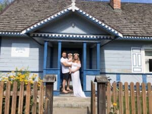 A man, woman, and child stand together in front of a blue wooden house with a picket fence and yellow flowers, capturing the charm of Poland summer.