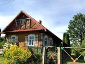 A wooden house with decorative trim, a red roof, and a fenced garden of blooming yellow flowers sits under a clear blue sky&mdash;a charming sight in Podlaskie, Poland, and one of the delightful things to do in the region.