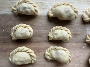 Seven uncooked sauerkraut pierogi with braided edges are arranged on a wooden surface, showcasing Babcia&rsquo;s secret recipe and an unmistakably authentic touch, ready to be cooked.