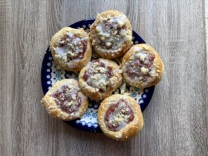 A plate of six round Drożdż&oacute;wki&mdash;Polish Apple Yeast Buns&mdash;with fruit filling, glaze, and crumb topping, arranged on a patterned blue dish on a wooden surface. An easy recipe for a classic treat.