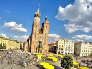 Aerial view of a busy city square with a large historic church, yellow market umbrellas, and people gathered under a partly cloudy sky.